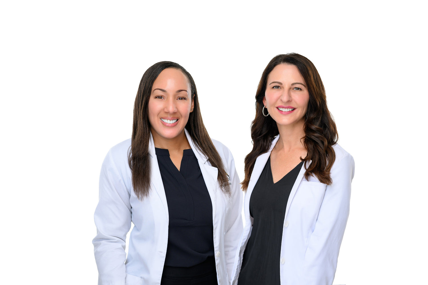 The image shows two smiling women wearing white lab coats over black tops. They are standing side by side against a plain white background. Both have long, dark hair. The woman on the left has straight hair, and the woman on the right has wavy hair. They are professional and approachable skincare experts.